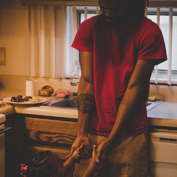 A man in a red shirt smiles while holding hands with a young girl with braided hair in a kitchen, standing near a counter with food. Warm lighting creates a cozy, intimate atmosphere.