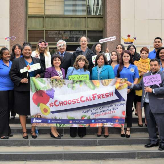 A diverse group of people stands on steps outside a building, smiling and holding signs promoting CalFresh and healthy eating. A large banner in front reads “#ChooseCalFresh” with application information.
