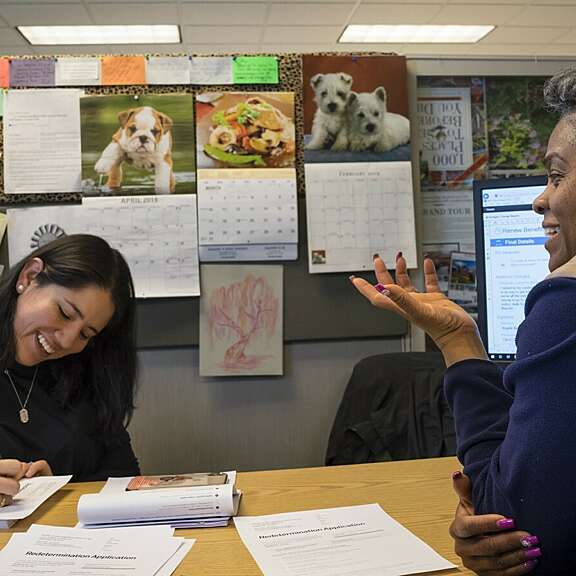 Three women sit at an office desk covered with papers; one is writing, another is speaking and gesturing, and the third listens. The background shows a bulletin board with calendars, photos of animals, and colorful notes.