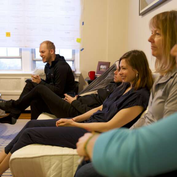 A group of people sit closely together on a couch and chairs in a brightly lit room, engaging in conversation, with a large paper schedule and windows visible in the background.