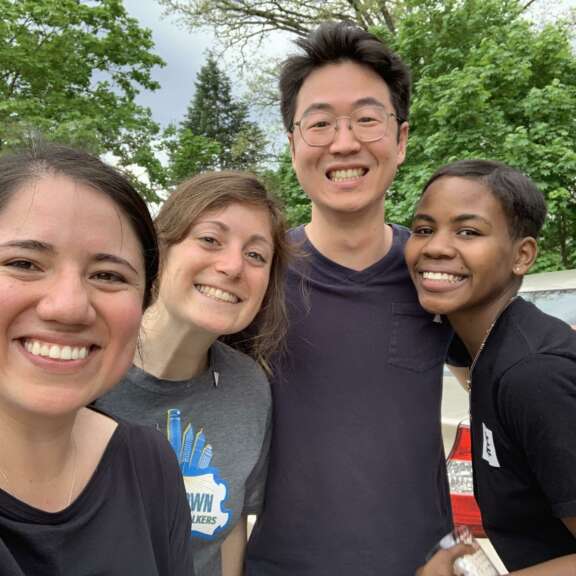 Four people stand close together outdoors, smiling at the camera. Trees and a parked car are visible in the background. Everyone appears happy and relaxed.