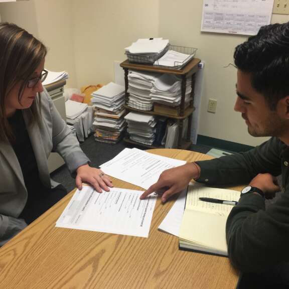Two people sit at a desk in an office, reviewing and discussing documents. One person points at a paper on the desk, while the other listens and takes notes. Stacks of paper and office supplies are visible in the background.