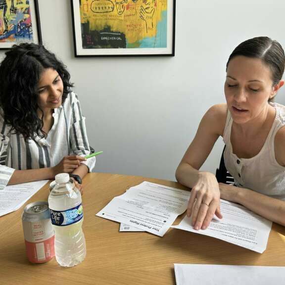 Two women sit at a round table reviewing documents together. One gestures to the papers while the other listens. A can, a water bottle, and more papers are on the table. Colorful artwork hangs on the wall behind them.