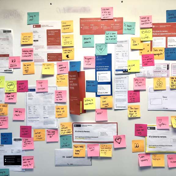 A whiteboard covered with colorful sticky notes, papers, and printouts. The notes contain handwritten ideas, sketches, and symbols, indicating brainstorming or project planning.