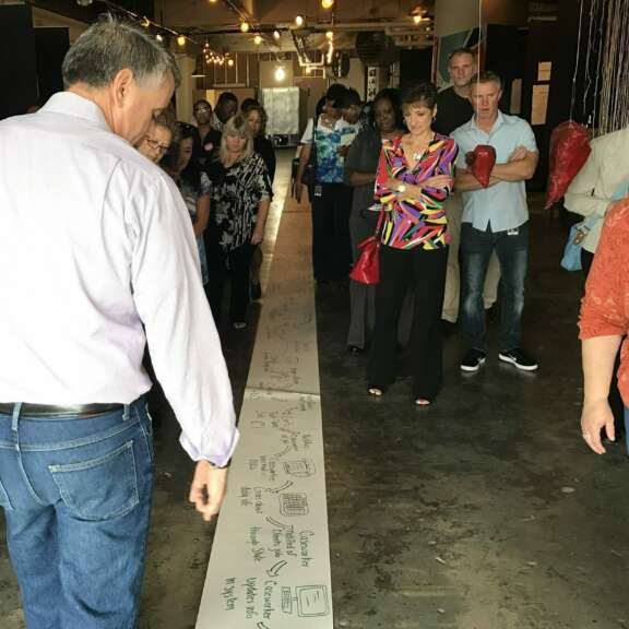 A group of people stands around a long sheet of paper on the floor covered with handwritten notes and drawings, observing and discussing its contents in an indoor, well-lit space.