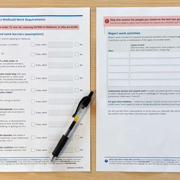 Two Medicaid forms and a black pen on a wooden table. The left form is about Medicaid work requirements; the right form is for reporting work activities. Both have checkboxes and sections to fill out.