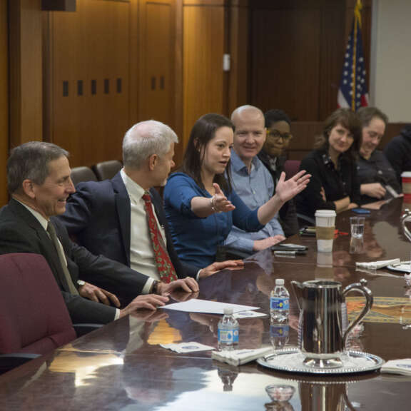 A group of people, including men and women, sit around a large conference table having a discussion. One woman gestures with her hands as she speaks. Papers, water bottles, and pitchers are on the table. An American flag is in the background.