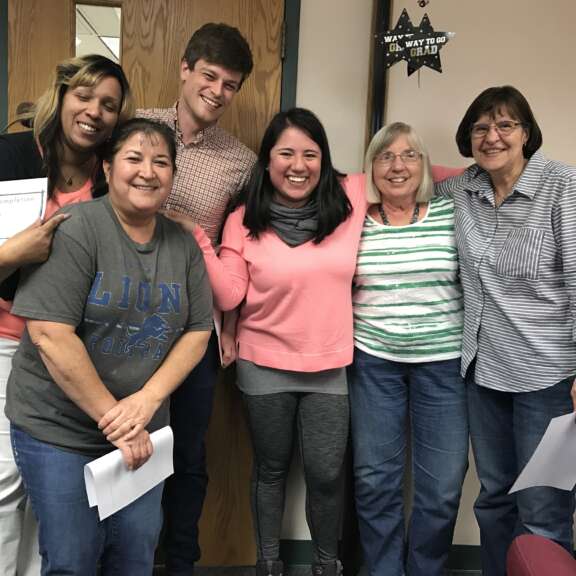 Six people stand together smiling in a room, holding certificates and papers. They appear happy and close, with arms around each other. A “Way to Go, Grad” sign and wood door are visible in the background.