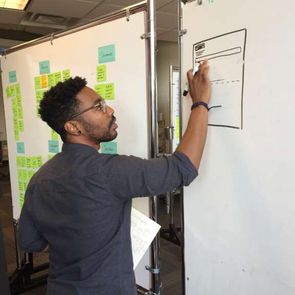 A man wearing glasses draws a wireframe on a whiteboard in an office, with colorful sticky notes and project plans visible in the background. He holds a paper in his left hand while working.
