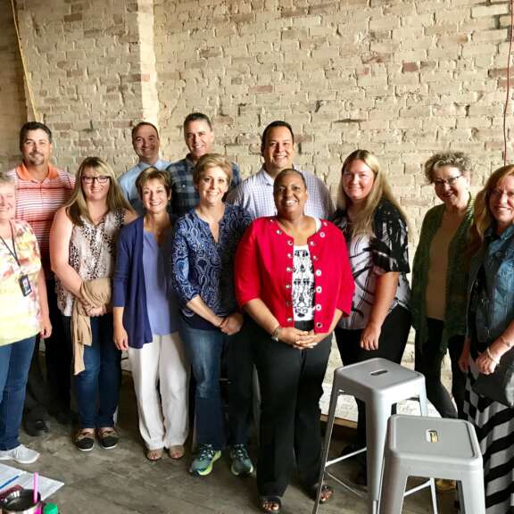 Thirteen people stand together indoors in front of a brick wall, smiling at the camera. There are stools nearby and some papers and bags on the floor. The group appears to be informal and friendly.