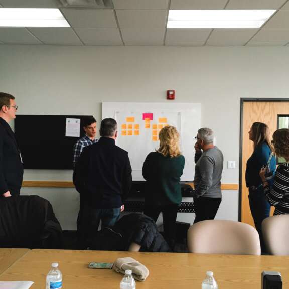 Six people stand in front of a whiteboard covered with sticky notes, discussing ideas in a conference room. Some sit at a table with water bottles, while others face the board, appearing engaged in conversation.