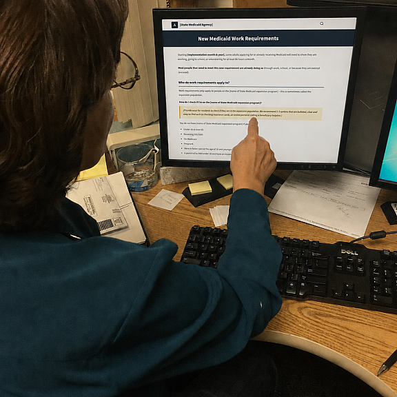A person wearing glasses sits at a desk, pointing at a computer screen displaying a New Medicaid Work Requirements webpage. Papers, a keyboard, and a phone are also visible on the desk.