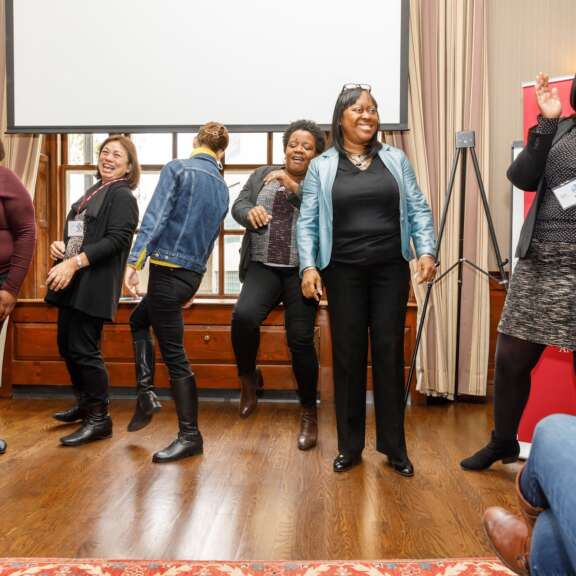 Six women stand together indoors, laughing and smiling, some clapping or dancing. They appear to be enjoying a lively moment during an event. A red banner and a projection screen are visible in the background.