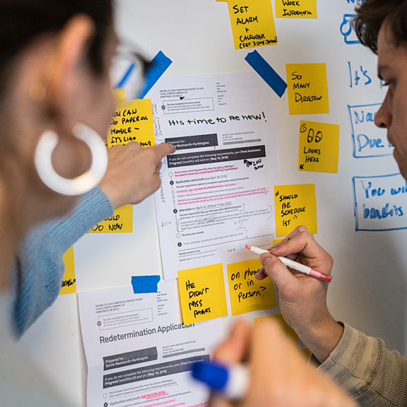 Two people review and annotate printed documents taped to a whiteboard, which is also covered with yellow sticky notes containing handwritten reminders and questions. One person points at the papers while the other writes with a marker.