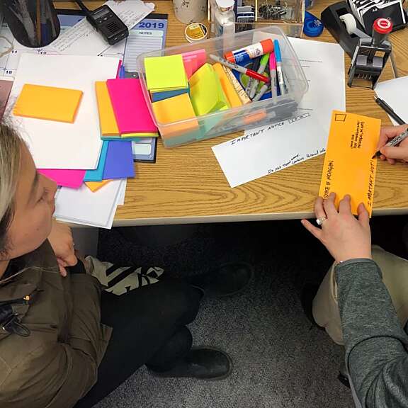Two people sit at a desk covered with colorful sticky notes, pens, markers, and office supplies. One person is writing on an orange envelope while the other observes. Papers and folders are scattered on the desk.