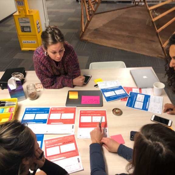 Four women sit around a table covered with colorful papers, notebooks, and pens, engaged in a collaborative discussion or planning session in a modern office setting.