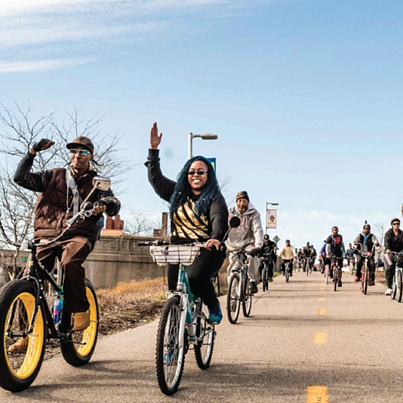 A group of people ride bicycles down a paved path on a sunny day, smiling and waving at the camera. Some trees and a bridge are visible in the background.