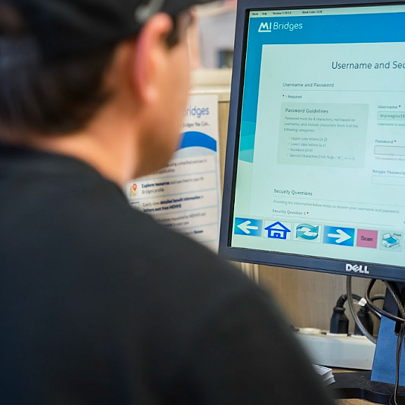 A person in a black cap and shirt sits at a desk using a computer, which displays a login page for a website called MI Bridges.
