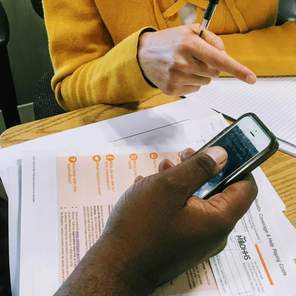 Two people sit at a table with paperwork, one holding a phone and the other pointing with a pen. The documents include forms labeled MDHHS. The setting appears to be an office or meeting room.