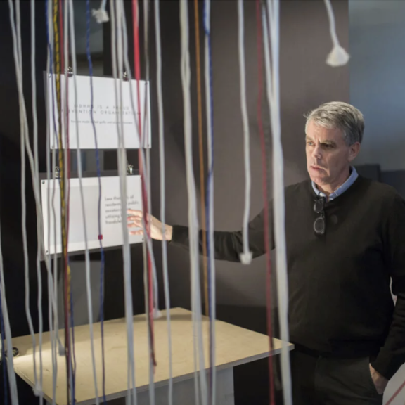 A man stands in a room, facing a display of hanging, colorful strings and reading information panels. He is dressed in a black sweater with glasses hanging from the collar. The room is dimly lit.