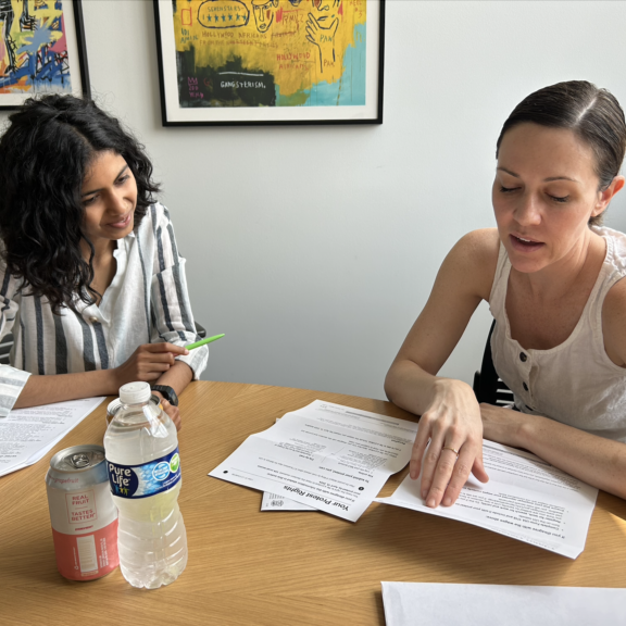 Two women sit at a table reviewing papers together. One points at a document while the other listens and holds a pen. On the table are drinks, including bottled water and a can. Colorful art hangs on the wall behind them.