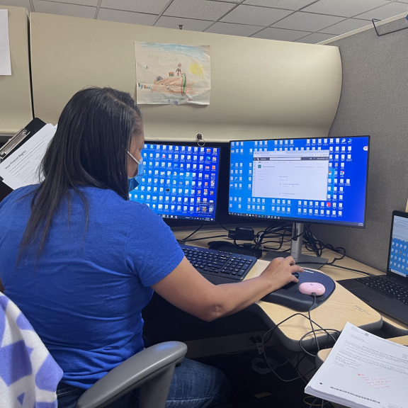 A woman wearing a blue shirt and face mask works at a desk with three computer monitors and a laptop, surrounded by papers and office supplies in a cubicle.