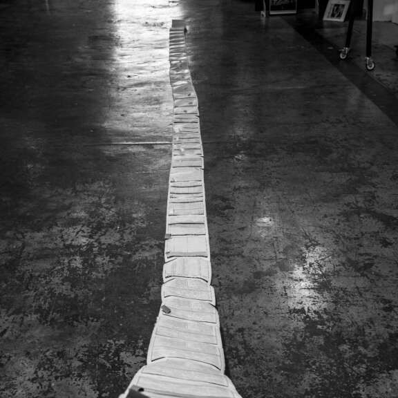 A long line of papers taped together is rolled out across a concrete floor, stretching into the distance in a dimly lit room. The image is in black and white.