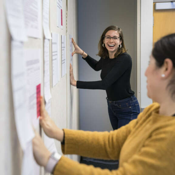 Two women are smiling and talking while pinning papers to a wall in an office or classroom setting. One woman is in the foreground wearing a yellow sweater, and the other is in the background wearing glasses and a black top.