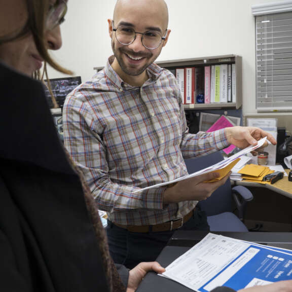 A man with glasses and a plaid shirt smiles while handing papers to a woman in a coat. They are standing in an office with shelves and a desk in the background.