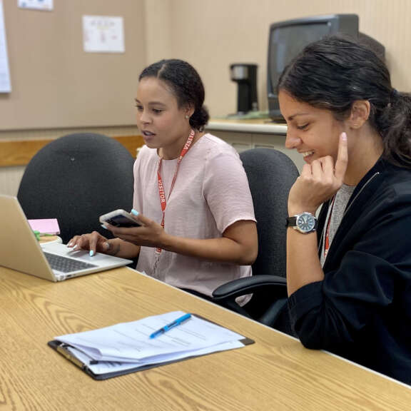 Two women sit at a conference table with a laptop and paperwork. One woman types on the laptop while the other looks at her phone and talks. Both appear focused and engaged in discussion.