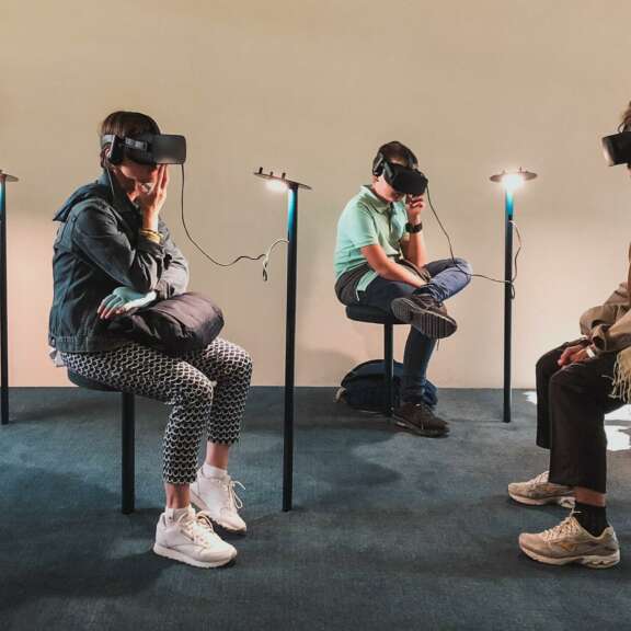 Four people are seated on stools in a room, each wearing virtual reality headsets and headphones, immersed in their experiences. The room is plain with light-colored walls and minimal lighting.