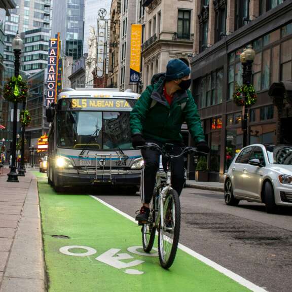 A cyclist rides in a green bike lane on a city street alongside cars and a bus. Pedestrians walk on the sidewalk, and buildings with shops and signs line both sides of the street.