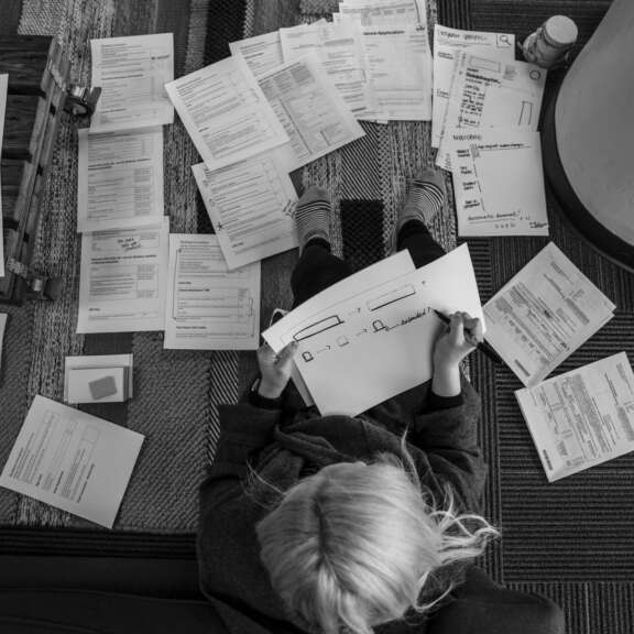 A person with light hair sits on the floor, surrounded by scattered papers and documents, reviewing forms and notes. Their back is to the camera and they are holding a sheet with diagrams. The setting appears to be an office.