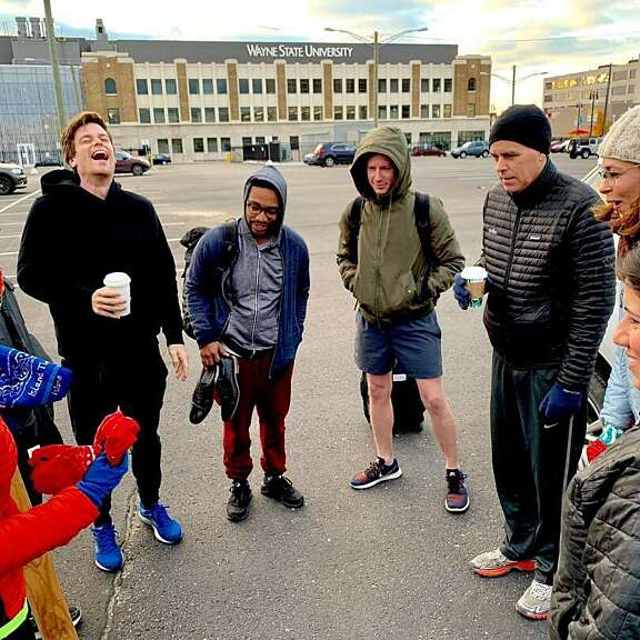 A group of eight people, dressed warmly, stand in a circle and talk in a parking lot. Some are holding drinks and laughing. Wayne State University is visible in the background.