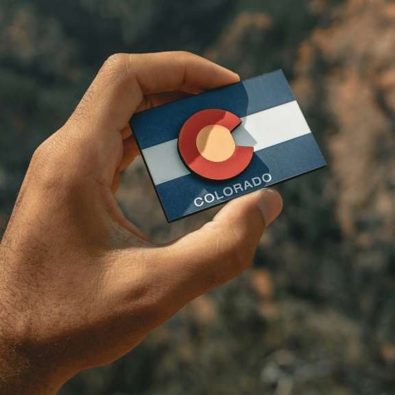 A hand holds a small card featuring the Colorado state flag against a blurred outdoor background of rocky terrain and greenery.