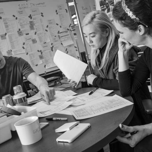 Three people sit around a table covered with papers, pens, and coffee cups, discussing documents. Behind them is a whiteboard filled with notes and diagrams. The image is in black and white.