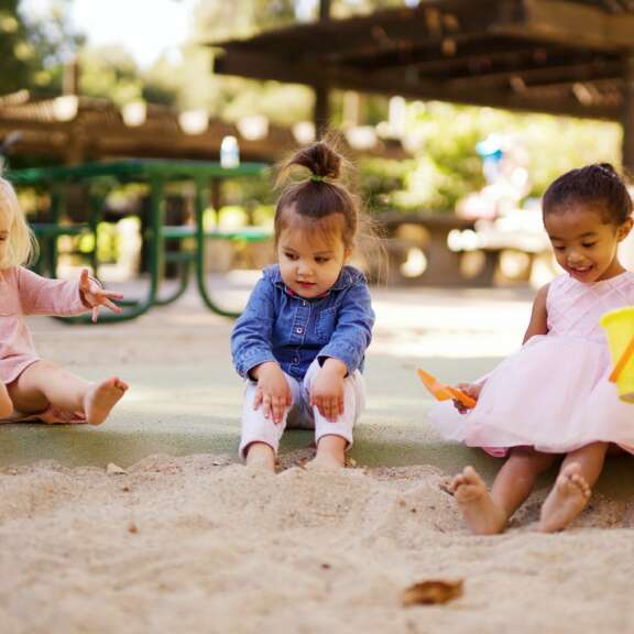 Three young children sit together in a sandbox outside. Two girls wear pink dresses, and one wears a blue denim shirt. They smile and play with sand, using a yellow bucket and orange shovel. Trees and picnic tables are in the background.