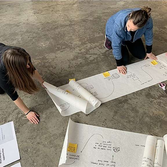 Two people kneel on a concrete floor, unrolling and reviewing a long sheet of paper with diagrams, notes, and sticky notes. Various stationery items are scattered nearby.