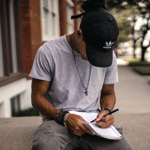 A person wearing a black Adidas cap and gray t-shirt sits outdoors, looking down and writing in a notebook resting on their lap. Trees and a building are visible in the background.