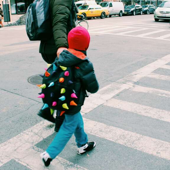 Child wearing a colorful spiked backpack and red hat walks across a city crosswalk, holding hands with an adult. Cars and taxis are visible in the background.
