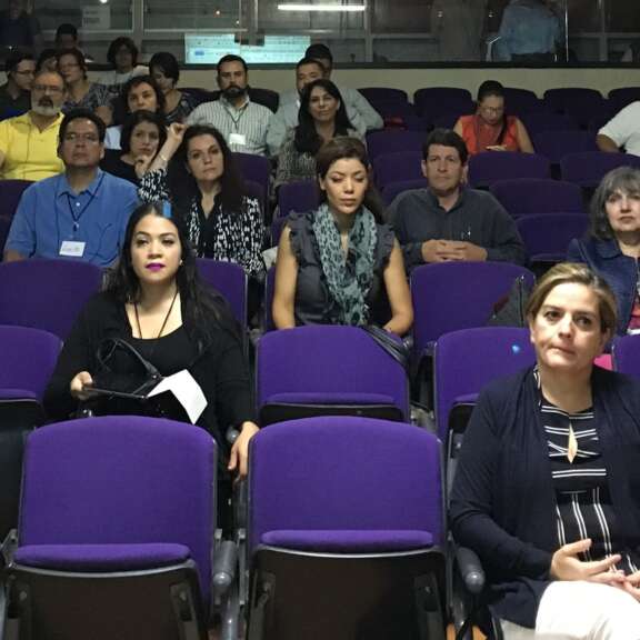 A diverse group of people sit in purple auditorium chairs, facing forward and attentively watching a presentation or event. Some hold papers or devices, while others sit with hands folded.