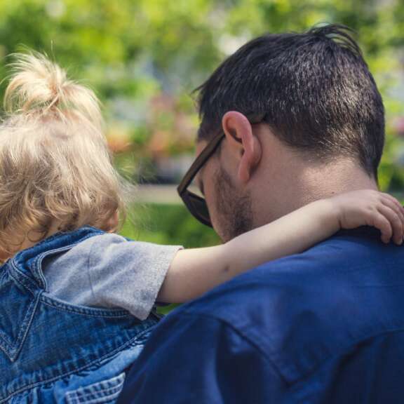 A man with dark hair and glasses carries a young child with blonde hair in a ponytail, outdoors. The child’s arm rests on the mans shoulder, and both are seen from behind against a blurred green background.