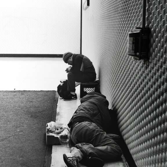 Two people are in an indoor corridor; one is sitting against the wall with a hood, looking down, while the other is lying on the ground covered by a coat. The scene is in black and white.