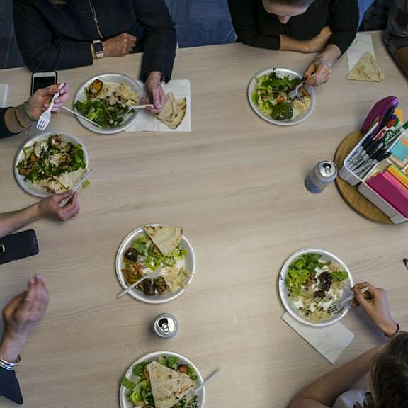 A group of people sit around a light wooden table eating salads and flatbread from plates, with drinks and napkins. Office supplies are in the center of the table. The photo is taken from above.