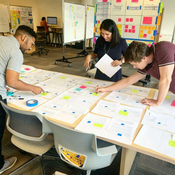 Three people are standing around a large table covered with papers and sticky notes, collaborating and making notes. Whiteboards with more notes and diagrams are visible in the background, suggesting a team brainstorming session.