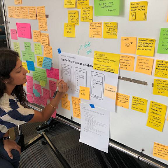 A woman kneels by a whiteboard covered with colorful sticky notes and sketches of mobile app screens, writing on a note as part of a brainstorming or design session.