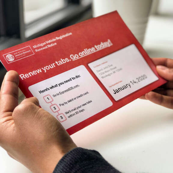 A person holds a red Michigan Vehicle Registration Renewal Notice envelope, which lists steps to renew tabs online and displays a due date of January 14, 2020.