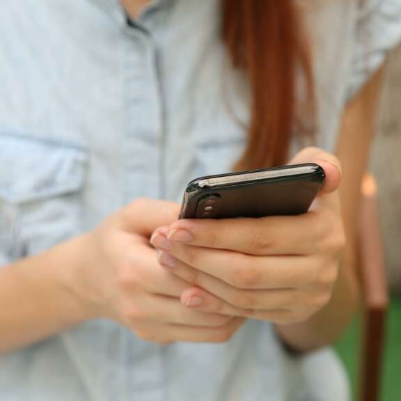 A person wearing a light blue, short-sleeved shirt is holding and using a smartphone with both hands. The background is blurred, showing some greenery and indoor elements.