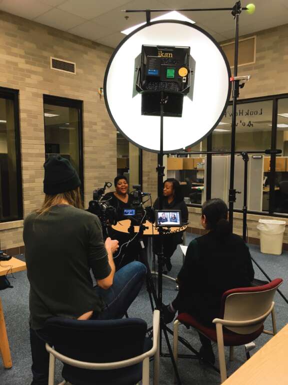 A behind-the-scenes view of a video interview setup, with two women seated at a table under bright lighting, while two crew members operate a camera and monitor in a room with bookshelves and glass windows.