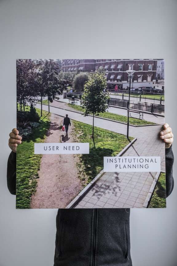 A person holds a poster showing a park with a paved walkway and a dirt path cutting across the grass. Labels read USER NEED on the dirt path and INSTITUTIONAL PLANNING on the paved walkway.